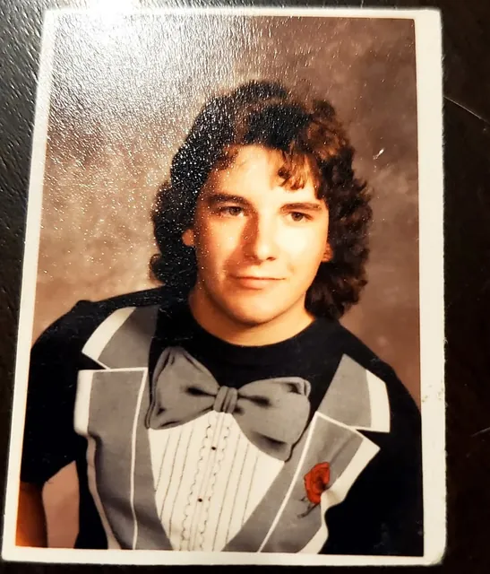 John's 1989 senior photo: big curly hair and a tuxedo T-shirt